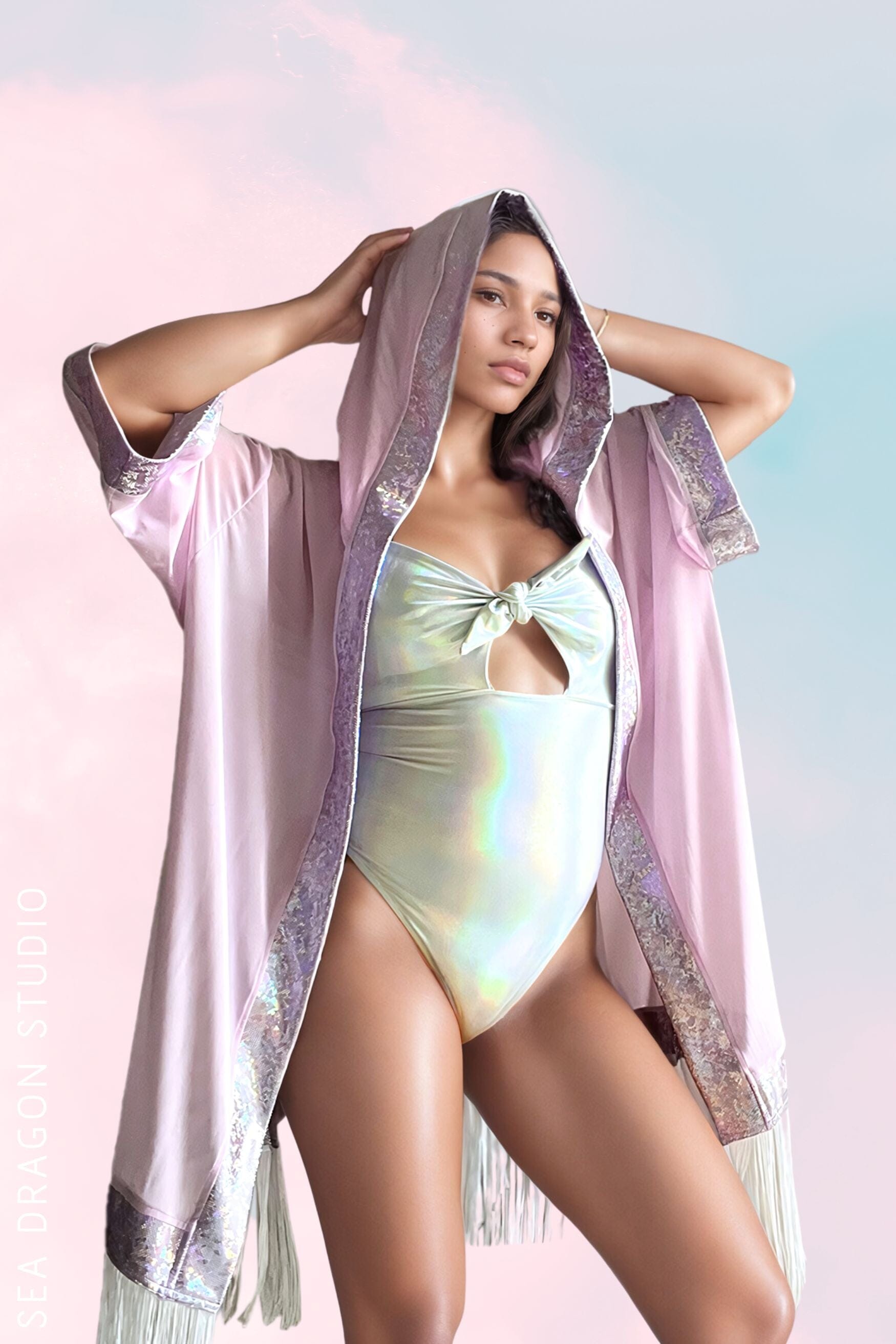 Model wearing a pastel pink hooded kimono with holographic trim and fringe over a holographic light mint one-piece bodysuit. The outfit is styled for a festival or beachwear, highlighting a chic and ethereal aesthetic.