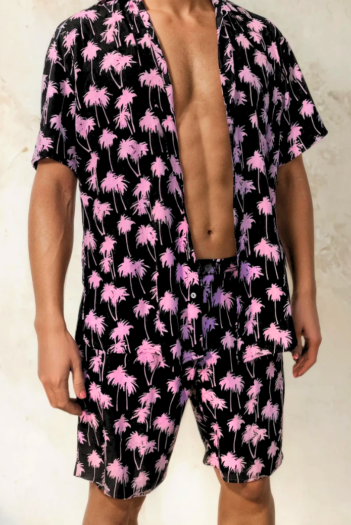 Model wearing the Malibu Barbie Palm Party rayon shorts in pink palm print, standing against a light textured wall.