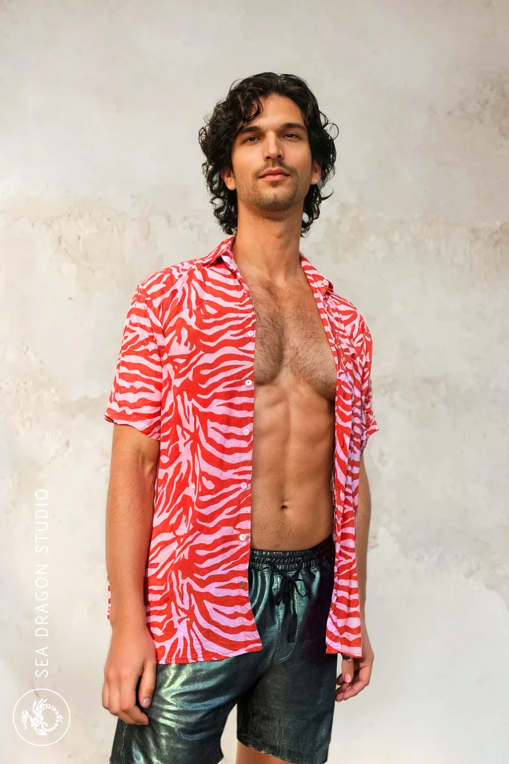 Model wearing the Coastal Mirage Heatwave Shirt in warm abstract print, photographed against a textured wall.