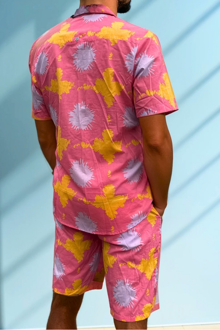Back view of the Tropic Drift men’s cotton shorts in pink sunshine print, photographed against a blue wall.