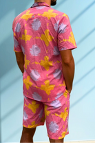 Back view of the Tropic Drift men’s cotton shorts in pink sunshine print, photographed against a blue wall.