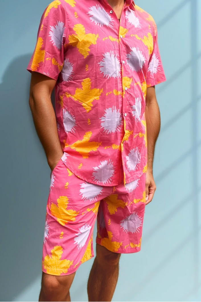 Model wearing the Tropic Drift pink cotton shorts, standing beside a blue wall in soft sunlight.